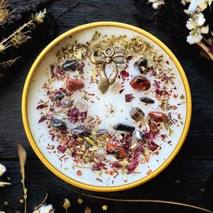 May include: A round candle in a yellow container featuring a white wax surface topped with scattered dried flower petals, small green herbs, various polished dark and reddish-brown stones, and a central metallic bow-shaped charm.