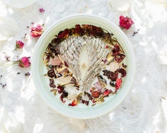 May include: A round white candle in a light-colored container, topped with a silver-toned metal angel wing charm, dried red rose petals, and various small crystals embedded in the wax around a central cotton wick.