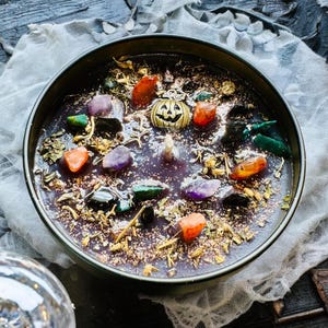 Peut inclure: Un r&eacute;cipient &agrave; bougie noir rond rempli de cire fonc&eacute;e, surmont&eacute; de pierres pr&eacute;cieuses polies color&eacute;es, d'herbes s&eacute;ch&eacute;es, de paillettes dor&eacute;es et d'une petite breloque citrouille en m&eacute;tal dor&eacute; centr&eacute;e autour d'une m&egrave;che en coton.
