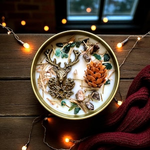 May include: A round, gold-colored candle with a deer head emblem, pine cone, and decorative stones. The candle is filled with white wax and surrounded by straw-like material. The candle sits on a wooden surface with string lights and a burgundy scarf.