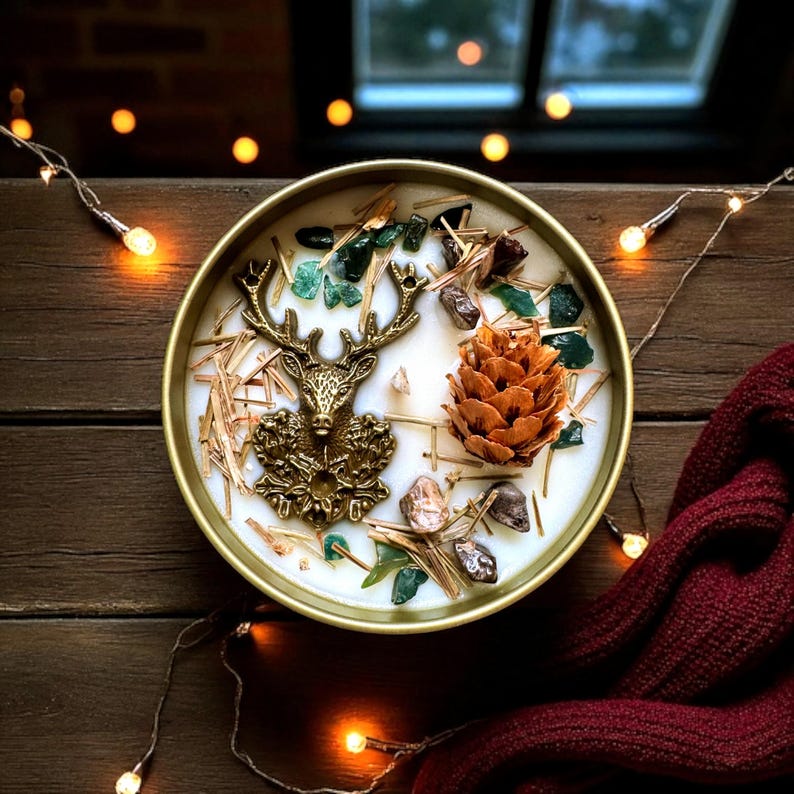 May include: A round, gold-colored candle with a deer head emblem, pine cone, and decorative stones. The candle is filled with white wax and surrounded by straw-like material. The candle sits on a wooden surface with string lights and a burgundy scarf.