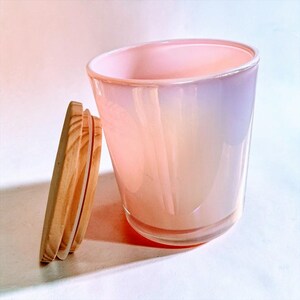 May include: A cylindrical glass candle vessel featuring an iridescent, pearlescent finish that shifts between soft pink and light purple tones. Beside the vessel stands a circular, light-coloured wooden lid with a visible grain and a silicone seal.