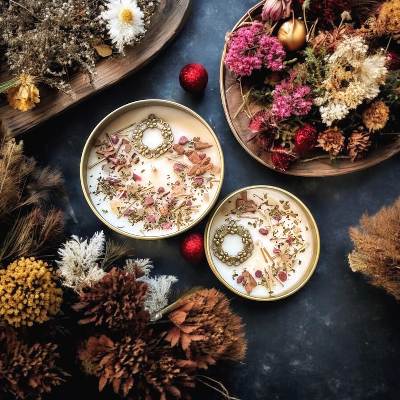 May include: Two round gold-toned candle tins filled with white wax, topped with dried herbs, small pink stone fragments, and a decorative gold-colored metal wreath charm. The candles are surrounded by various dried flowers and foliage.