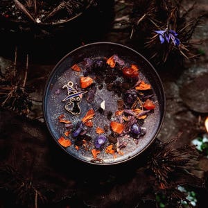 May include: A round black candle in a container topped with dark wax, silver glitter, purple and orange crystal fragments, dried flower petals, and a small metal charm shaped like a black cat wearing a purple and white striped witch hat.