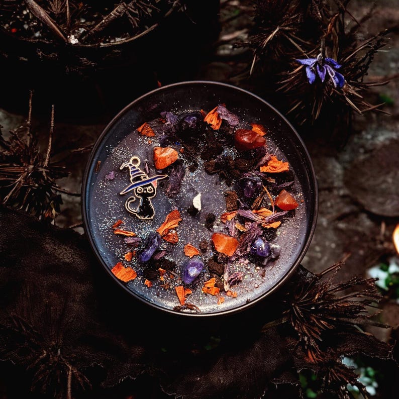 May include: A round black candle in a container topped with dark wax, silver glitter, purple and orange crystal fragments, dried flower petals, and a small metal charm shaped like a black cat wearing a purple and white striped witch hat.