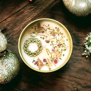 May include: A round gold-colored metal tin containing white wax, topped with a circular bronze-toned floral charm, small pink stones, and scattered dried botanical pieces. The candle is presented from a top-down perspective.