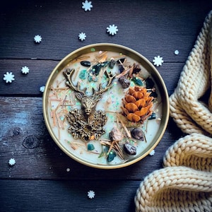 May include: A round candle in a gold-colored metal container. The candle is decorated with a bronze-colored deer, a pine cone, green stones, and small pieces of wood. The candle sits on a dark wooden surface with white snowflake decorations.
