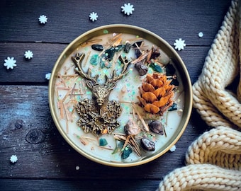 May include: A round candle in a gold-colored metal container. The candle is decorated with a bronze-colored deer, a pine cone, green stones, and small pieces of wood. The candle sits on a dark wooden surface with white snowflake decorations.