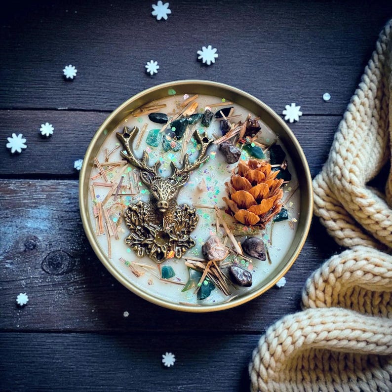 May include: A round candle in a gold-colored metal container. The candle is decorated with a bronze-colored deer, a pine cone, green stones, and small pieces of wood. The candle sits on a dark wooden surface with white snowflake decorations.