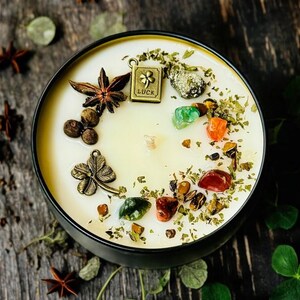 May include: A round black candle tin filled with cream-coloured wax, topped with a metal charm shaped like a book with the word LUCK and a four-leaf clover, a metal four-leaf clover charm, star anise, peppercorns, and various colourful gemstones.