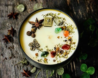 May include: A round black candle tin filled with cream-colored wax, topped with a metal charm shaped like a book with the word LUCK and a four-leaf clover, a metal four-leaf clover charm, star anise, peppercorns, and various colorful gemstones.