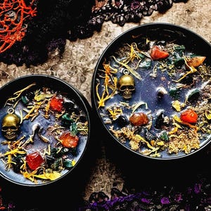 Puede incluir: Dos tarros de velas negras con dijes de calavera dorados y varios cristales y hierbas en su interior. Los tarros est&aacute;n sobre un fondo oscuro con encaje negro.