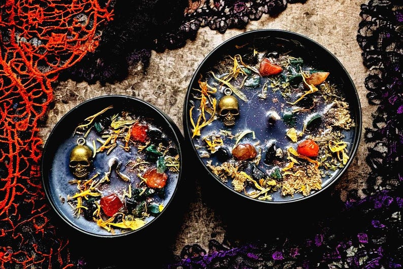 Puede incluir: Dos tarros de velas negras con dijes de calavera dorados y varios cristales y hierbas en su interior. Los tarros est&aacute;n sobre un fondo oscuro con encaje negro.
