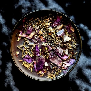 May include: A dark purple candle with a gold star charm and dried flowers and crystals on top. The candle is in a dark brown glass jar.