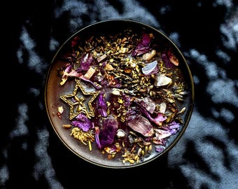 May include: A round black candle in a tin container topped with dried purple flower petals, small green herbs, clear and purple crystal stones, and a gold-toned metal star charm featuring a small figure inside the star shape.