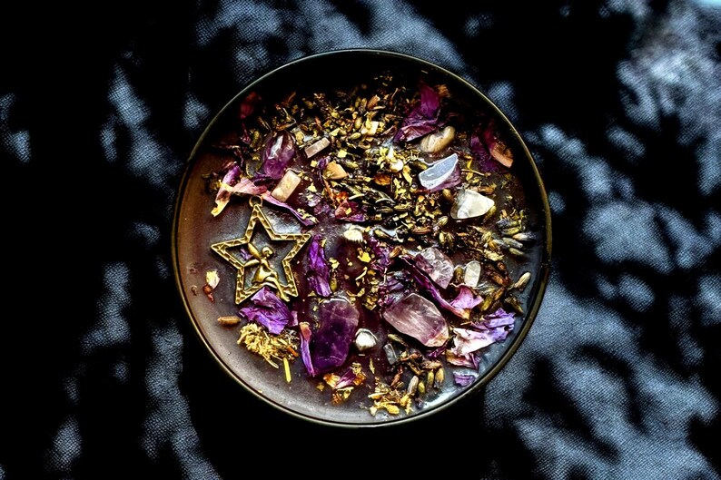 May include: A dark purple candle with a gold star charm and dried flowers and crystals on top. The candle is in a dark brown glass jar.