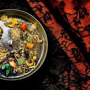 Puede incluir: Una vela negra con un dije de calavera dorada y varias hierbas y cristales incrustados en la cera. La vela est&aacute; en un cuenco negro y est&aacute; sentada sobre una tela de encaje negra y naranja.