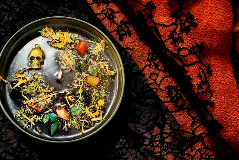 Puede incluir: Una vela negra con un dije de calavera dorada y varias hierbas y cristales incrustados en la cera. La vela est&aacute; en un cuenco negro y est&aacute; sentada sobre una tela de encaje negra y naranja.