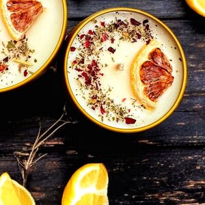May include: Two round gold-toned metal tins filled with off-white wax candles. Each candle features a single wooden wick, a dried orange slice, and a scattering of dried red flower petals and green herbs on the surface of the wax.