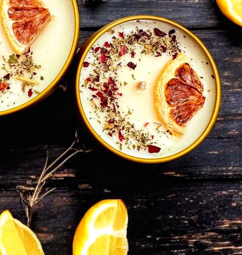 May include: Two round gold-toned metal tins filled with off-white wax candles. Each candle features a single wooden wick, a dried orange slice, and a scattering of dried red flower petals and green herbs on the surface of the wax.
