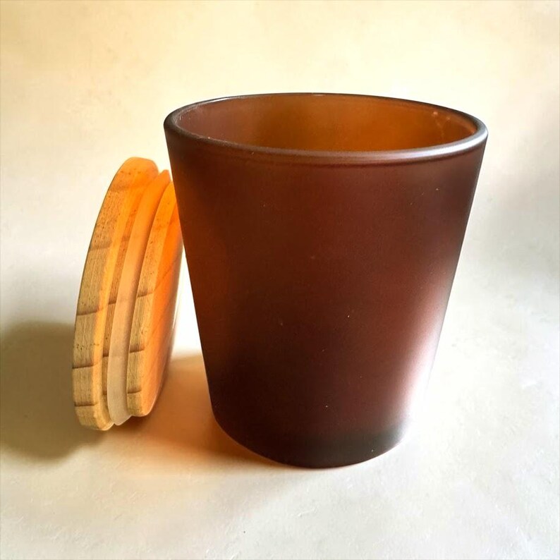 May include: A cylindrical candle jar made of dark brown frosted glass with a smooth, matte finish. Beside the jar stands a circular, light-coloured wooden lid featuring a visible grain pattern and a translucent silicone seal around its edge.