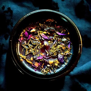 May include: A black bowl filled with a purple candle with gold glitter, dried flowers, and a gold star charm.