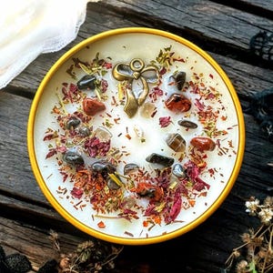 May include: A round yellow candle tin filled with white wax, topped with scattered dried flower petals, small dark and reddish-brown polished stones, and a central antique brass-coloured metal bow charm embedded in the surface of the wax.