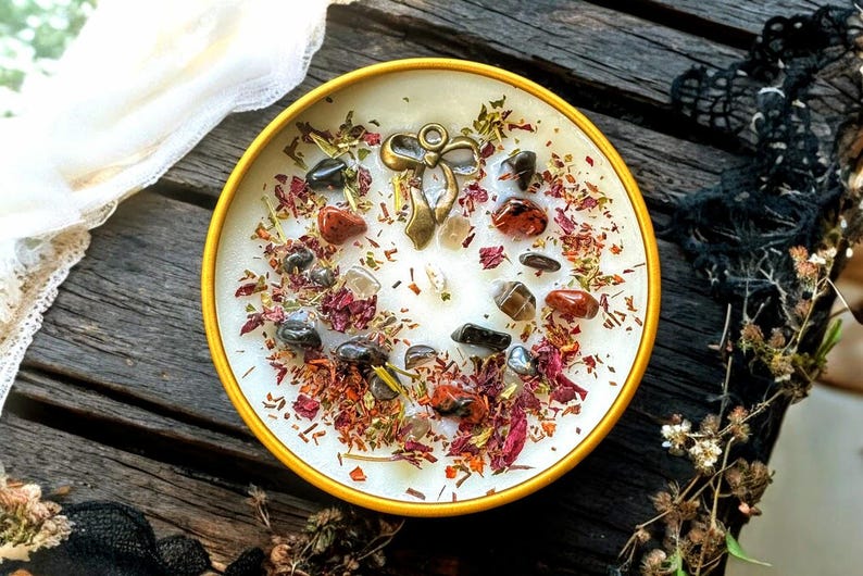 May include: A round yellow candle tin filled with white wax, topped with scattered dried flower petals, small dark and reddish-brown polished stones, and a central antique brass-coloured metal bow charm embedded in the surface of the wax.