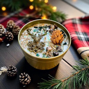 May include: A gold-colored candle tin filled with white wax, decorated with a bronze deer head, pine cone, and small stones. The candle is surrounded by pine needles and glitter. The background includes pine cones and a red plaid pattern.