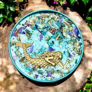May include: A teal blue round candle with a gold mermaid figurine embedded in the wax. The candle is surrounded by dried flowers and glitter.