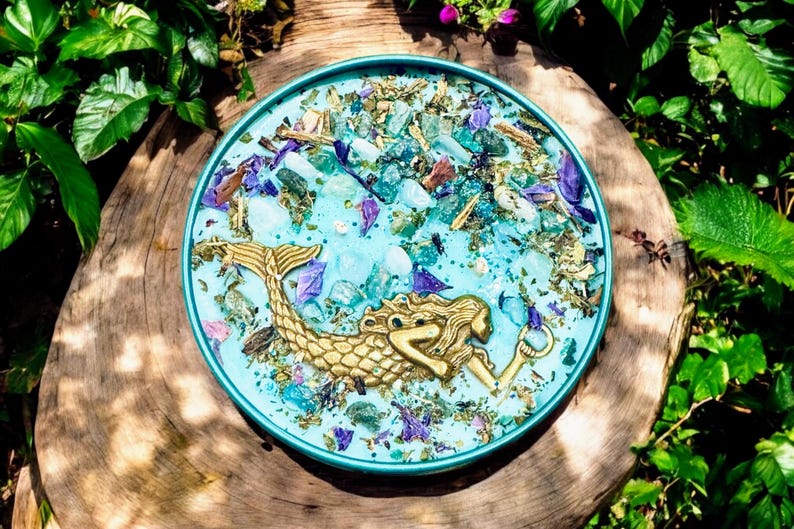 May include: A teal blue round candle with a gold mermaid figurine embedded in the wax. The candle is surrounded by dried flowers and glitter.