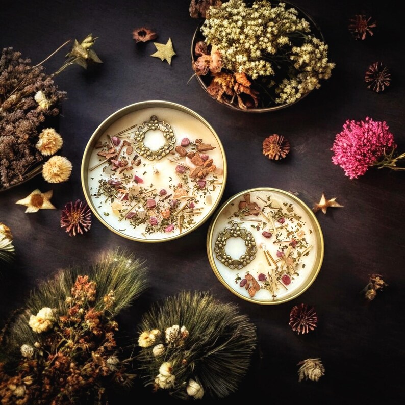May include: Two round gold-toned metal tins filled with white wax, each topped with a circular ornate bronze-colored metal charm, dried flower petals, small wood chips, and green herbs, surrounded by scattered dried flowers and pine needles.
