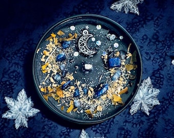 May include: A round black candle topped with blue tumbled stones, dried herbs, and a silver-toned crescent moon charm. The surface is decorated with small iridescent hexagonal sequins and a central wick, creating a dark, celestial-inspired appearance.