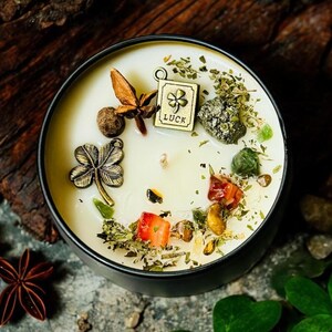 May include: A round black candle tin filled with cream-coloured wax, topped with a metal four-leaf clover charm, a square charm engraved with the word LUCK and a clover, star anise, dried herbs, small green stones, and red fruit pieces.