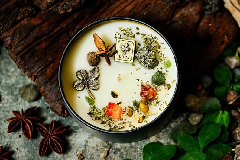 May include: A round black candle tin filled with cream-coloured wax, topped with a metal four-leaf clover charm, a square charm engraved with the word LUCK and a clover, star anise, dried herbs, small green stones, and red fruit pieces.