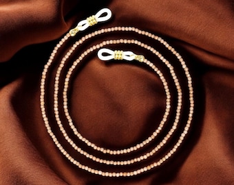 Könnte beinhalten: Eine dreifache Perlenkette für Brillen mit goldfarbenen Verschlüssen. Die Kette besteht aus kleinen, hellbraunen Perlen und liegt auf einer braunen Stoffoberfläche.