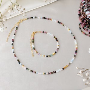 May include: A set of two necklaces made with colorful gemstone beads. The necklace is made with a variety of colors including pink, green, blue, and black. The necklace is made with small round beads and has a gold clasp.