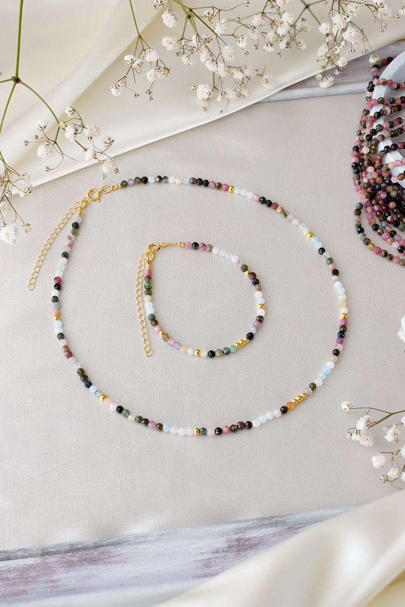 May include: A set of two necklaces made with colorful gemstone beads. The necklace is made with a variety of colors including pink, green, blue, and black. The necklace is made with small round beads and has a gold clasp.