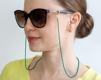 Könnte beinhalten: Eine Frau trägt eine dunkle Sonnenbrille mit einer grünen Perlenkette für Brillen. Die Kette ist um den Hals drapiert. Die Frau trägt einen gelben Pullover und goldene Ohrringe.