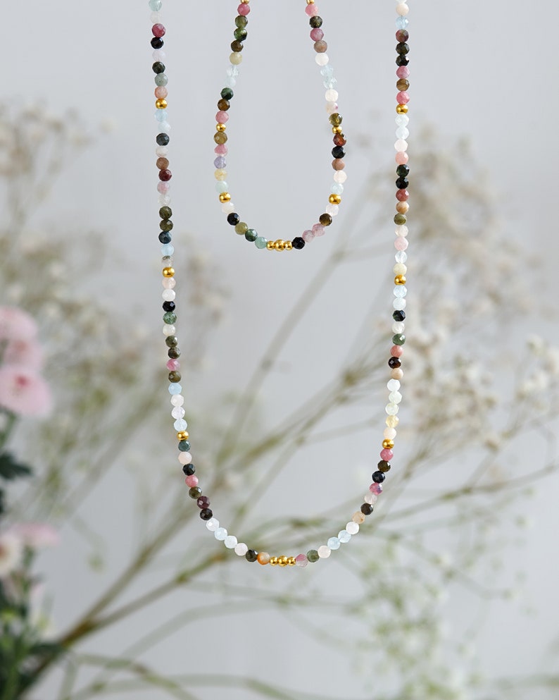 May include: A long and short necklace set with colorful gemstone beads and gold accents. The necklace is made of aquamarine, pink tourmaline, green tourmaline, black tourmaline, and other gemstones.