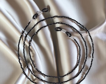 Könnte beinhalten: Schwarze Perlen-Brillenschnur mit silbernen Metallverschlüssen. Die Schnur besteht aus kleinen, runden Perlen und ist dazu bestimmt, Brillen um den Hals zu halten.