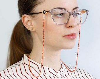 May include: A woman wearing glasses with a red beaded eyeglass chain. The chain is made of small, round beads and is attached to the temples of the glasses.