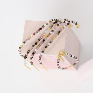 May include: A set of four multi-colored beaded bracelets with gold accents. The bracelets are layered on a pink surface.
