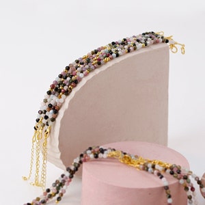 May include: Multi-colored gemstone beaded bracelets with gold chain accents. The bracelets are layered on top of each other and displayed on pink surfaces.