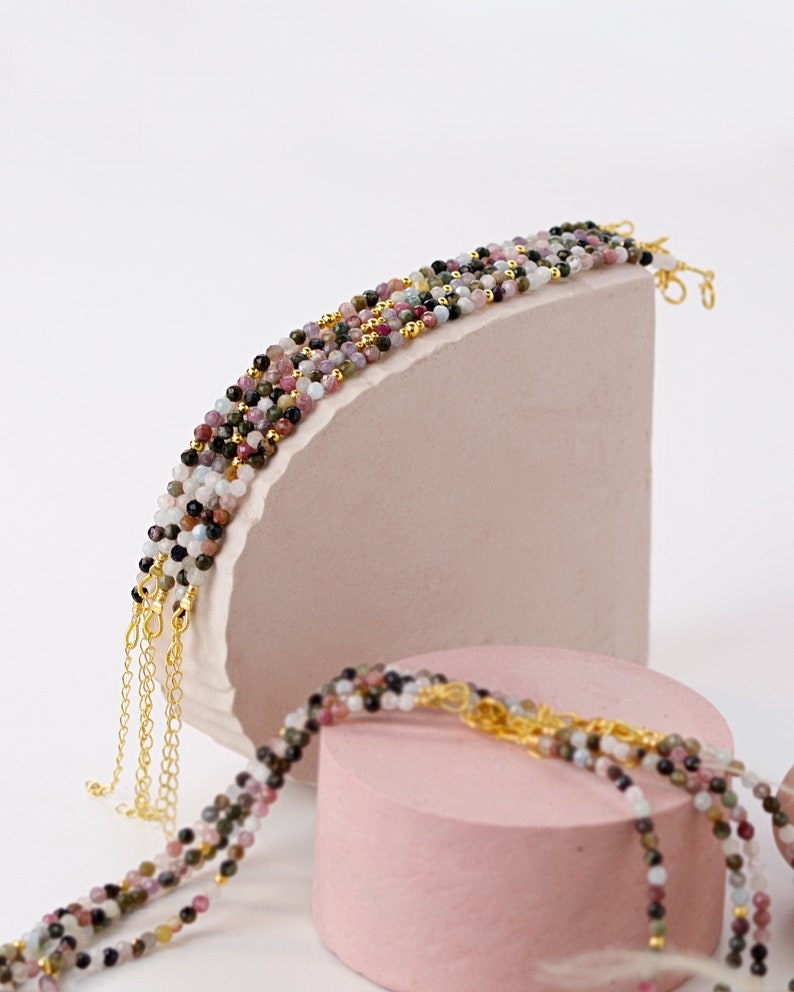 May include: Multi-colored gemstone beaded bracelets with gold chain accents. The bracelets are layered on top of each other and displayed on pink surfaces.