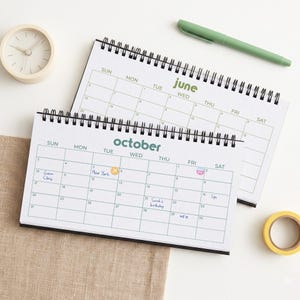 May include: Two spiral-bound desk calendars with white pages and black wire binding. One calendar shows the month of June in green text, and the other shows October in teal text with handwritten notes and stickers on the grid.
