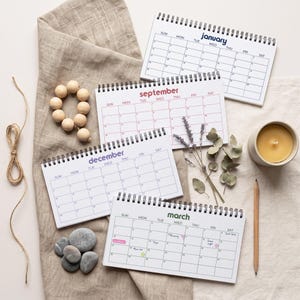 May include: Four spiral-bound desk calendars showing the months of January, September, December, and March. The calendars feature white pages with black grid lines and colored text for the month names. Also includes wooden beads, dried lavender, a candle, and a pencil.
