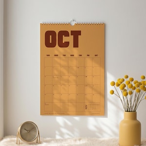 May include: A wall calendar with a mustard yellow background and dark brown text reading OCT at the top. The calendar features a grid for the month of October with thin black lines. A silver spiral binding holds the pages together at the top.