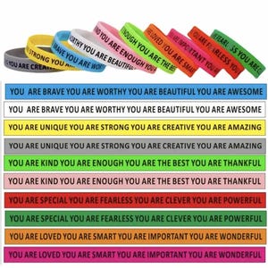 May include: A selection of colourful silicone wristbands featuring motivational phrases. The bands are in a range of colours, such as black, yellow, blue, white, pink, green, orange, red, and purple. The text on the bands includes phrases like "You are brave" and "You are enough."