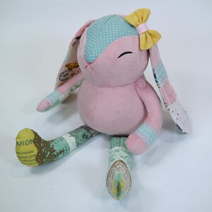 Puede incluir: Un conejito de peluche rosa hecho a mano con orejas de lunares azules y blancos, un lazo amarillo y patas de patchwork. El conejito est&aacute; sentado sobre un fondo blanco.